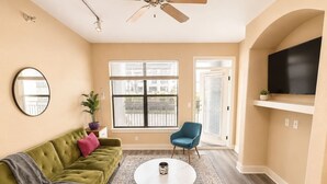 A living room with a green sofa, a blue chair, a round coffee table, and a TV on a wall. - Sleepover | Lavish 1BD/1BA + Pool - Little Rock (North Little Rock)