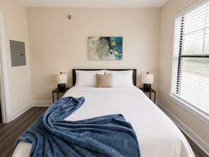 A bedroom with a large bed, two bedside tables with lamps, a painting on the wall, and a window with blinds.
