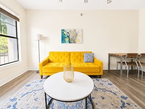A living room with a yellow sofa, a round coffee table, a patterned rug, a painting on the wall, and a dining area with a table and chairs.
