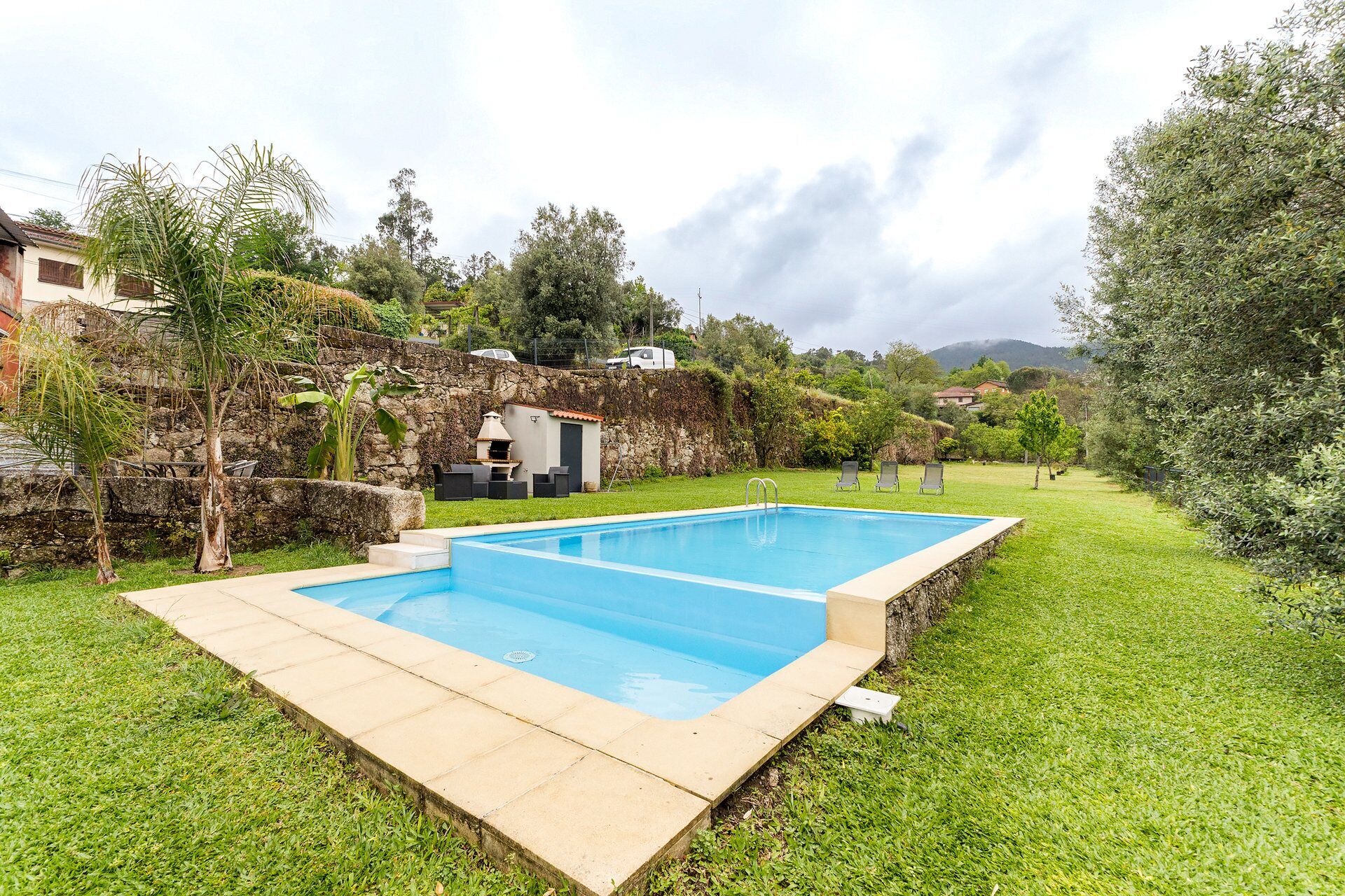 Une piscine rectangulaire bordée de pierres, entourée d’une pelouse et d’un mur en pierre.