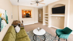 A living room with a green sofa, a round coffee table, a teal armchair, a flat-screen TV mounted on a wall, and a ceiling fan.