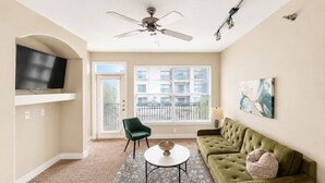 A living room with a green sofa, a round coffee table, a green chair, a flat-screen TV mounted on the wall, and a ceiling fan.