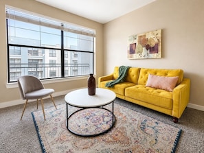 Una sala de estar con un sofá amarillo, una mesa de centro redonda, un sillón gris y un ventanal con persianas.