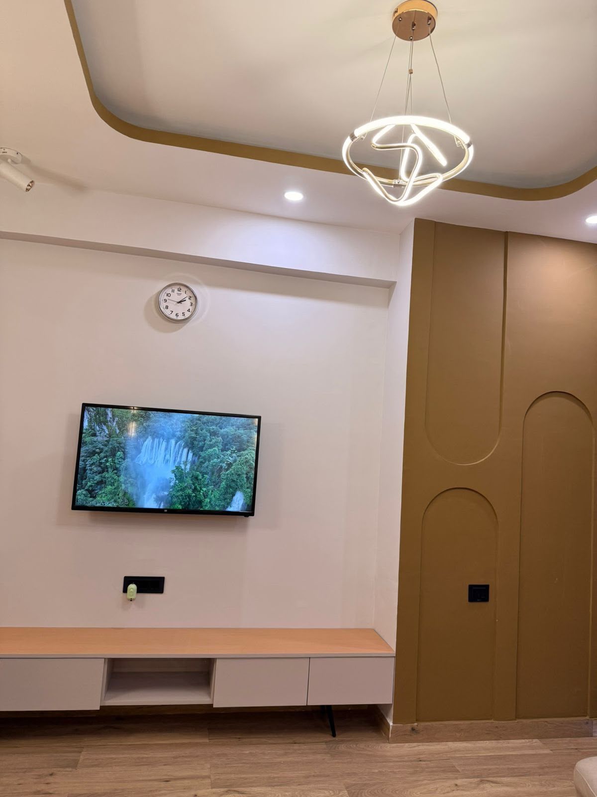 A modern living room with a flat-screen TV mounted on the wall, a wooden bench, a wall clock, and a unique ceiling light fixture.