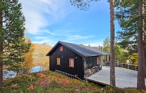 Una baita nera con un portico, circondata da alberi e da un lago, con una montagna sullo sfondo.