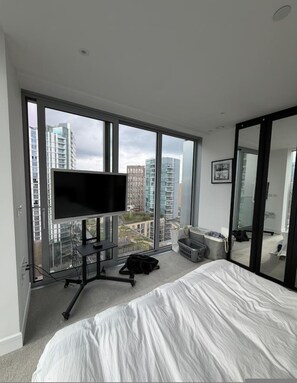 Ein modernes Hotelzimmer mit einem großen Bett, einem Fernseher auf einem Schrank, einem Schreibtisch und Blick auf Stadtgebäude durch große Fenster.