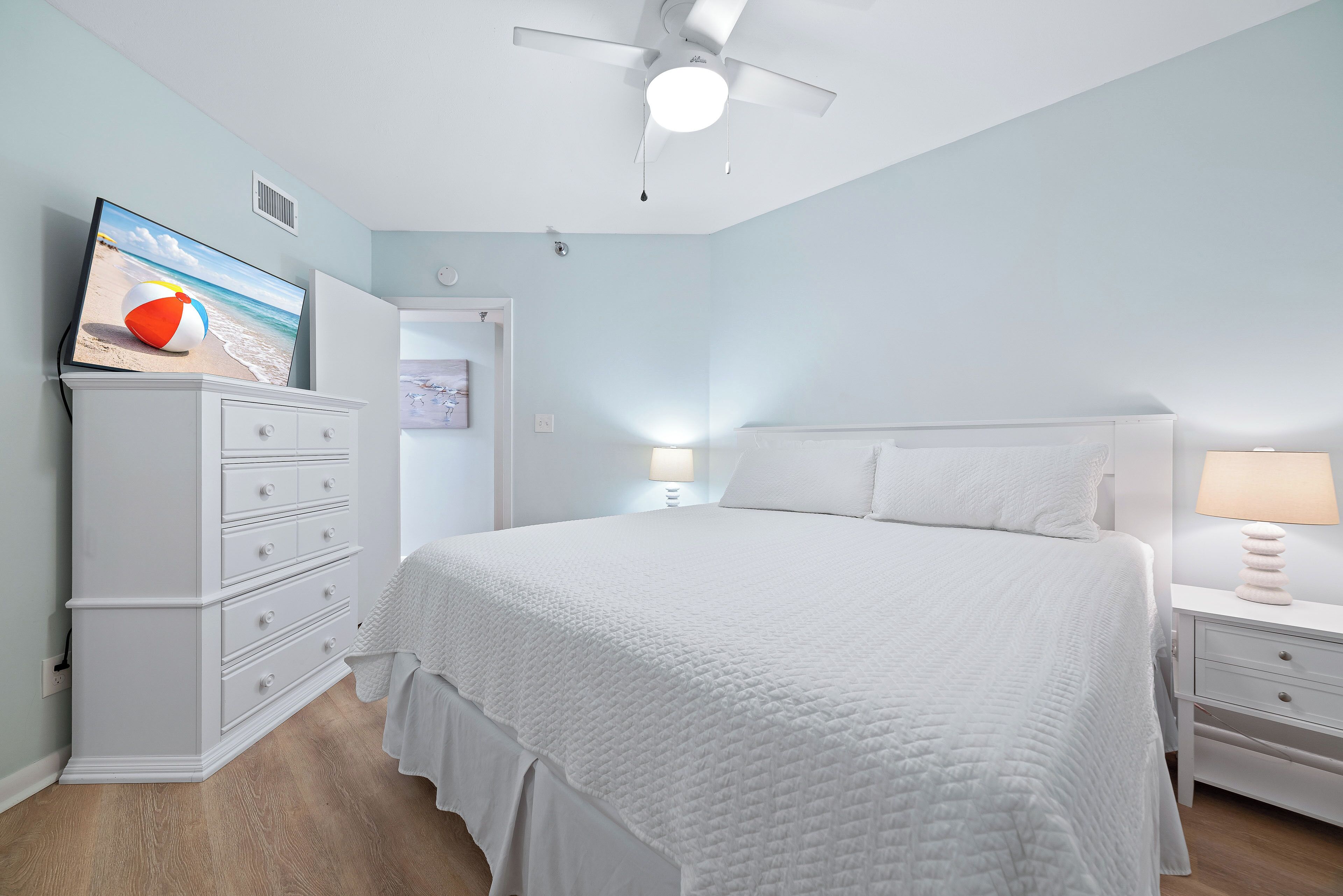 A bedroom with a bed, dresser, nightstand, lamp, ceiling fan, and a TV on the dresser.