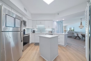 A modern kitchen with a central island, stainless steel appliances, and a dining area with a chandelier. - Beachfront Condo w/ Huge Balcony, Sleeps 11, Pools & Gulf Views | BEACHBALL PROPERTIES (Orange Beach)