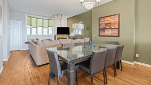 A modern living room with a glass dining table, grey chairs, a beige sofa, a flat-screen TV, and a chandelier. - Ultimate Slopeside Retreat – Sleeps 24 Across Two Spacious Townhomes (Silver Star Mountain)