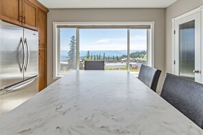 A kitchen with a large island, a stainless steel refrigerator, and a view through large windows.