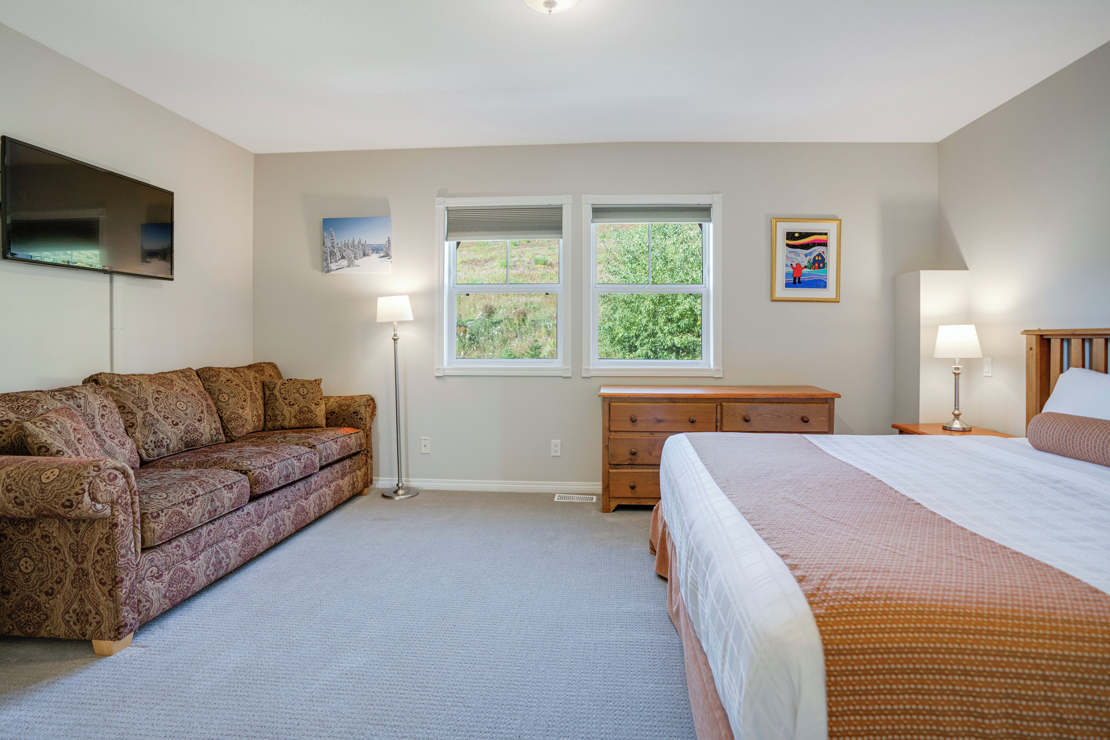 A bedroom with a bed, a sofa, a dresser, two windows, and a flat-screen TV.