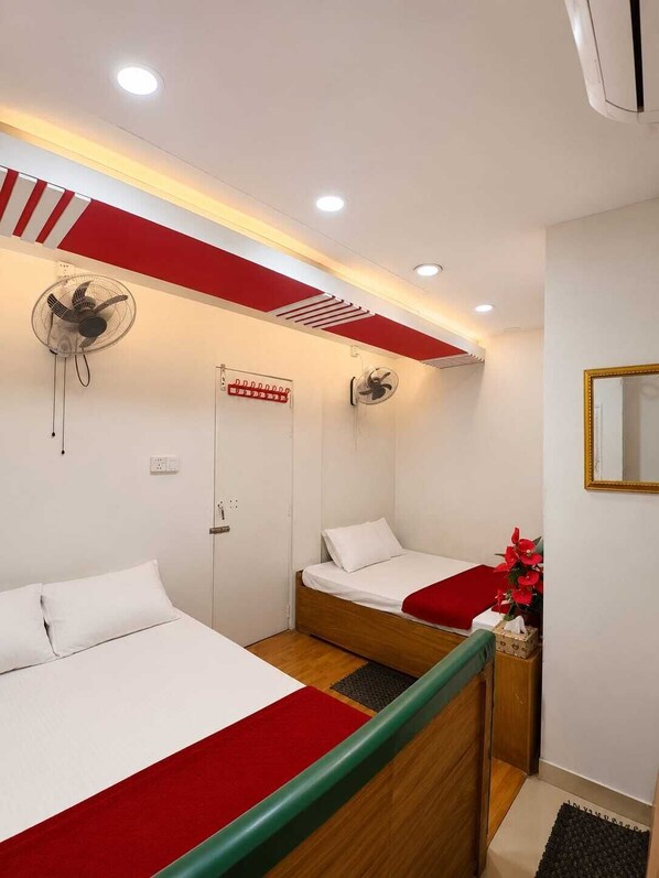 Una habitación de hotel con dos camas, techo a rayas rojas y blancas, un ventilador de pared, un espejo y una mesita con un jarrón de flores.