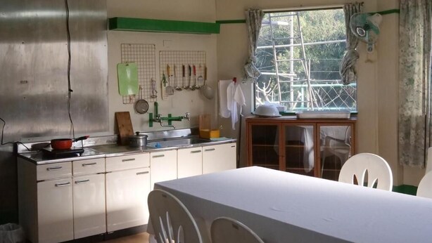 Una cocina con estufa, lavamanos y armarios, una mesa de comedor con sillas y una ventana con cortinas.