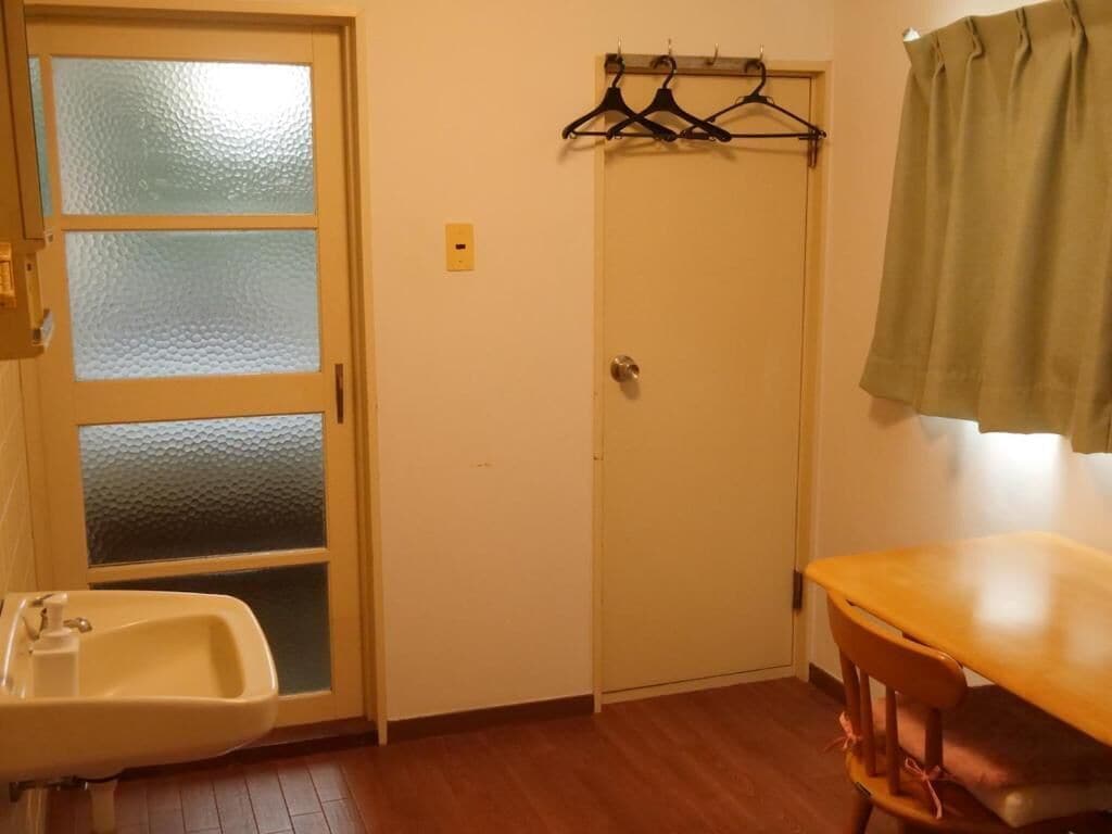 Quarto com piso de madeira, pia branca, porta com painéis de vidro fosco, cabideiro com ganchos e mesa de madeira com uma cadeira.