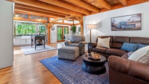 A spacious living area with a wooden ceiling, a brown leather sofa, a round coffee table, and a dining area with a stone wall and large windows. - Great Aspen Getaway | Luxury, Pet-Friendly Home with Game Room & Fire Pit! (Black Mountain)