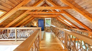 An attic room with wooden beams, a stone wall, a wooden railing, and a small table. - Great Aspen Getaway | Luxury, Pet-Friendly Home with Game Room & Fire Pit! (Black Mountain)