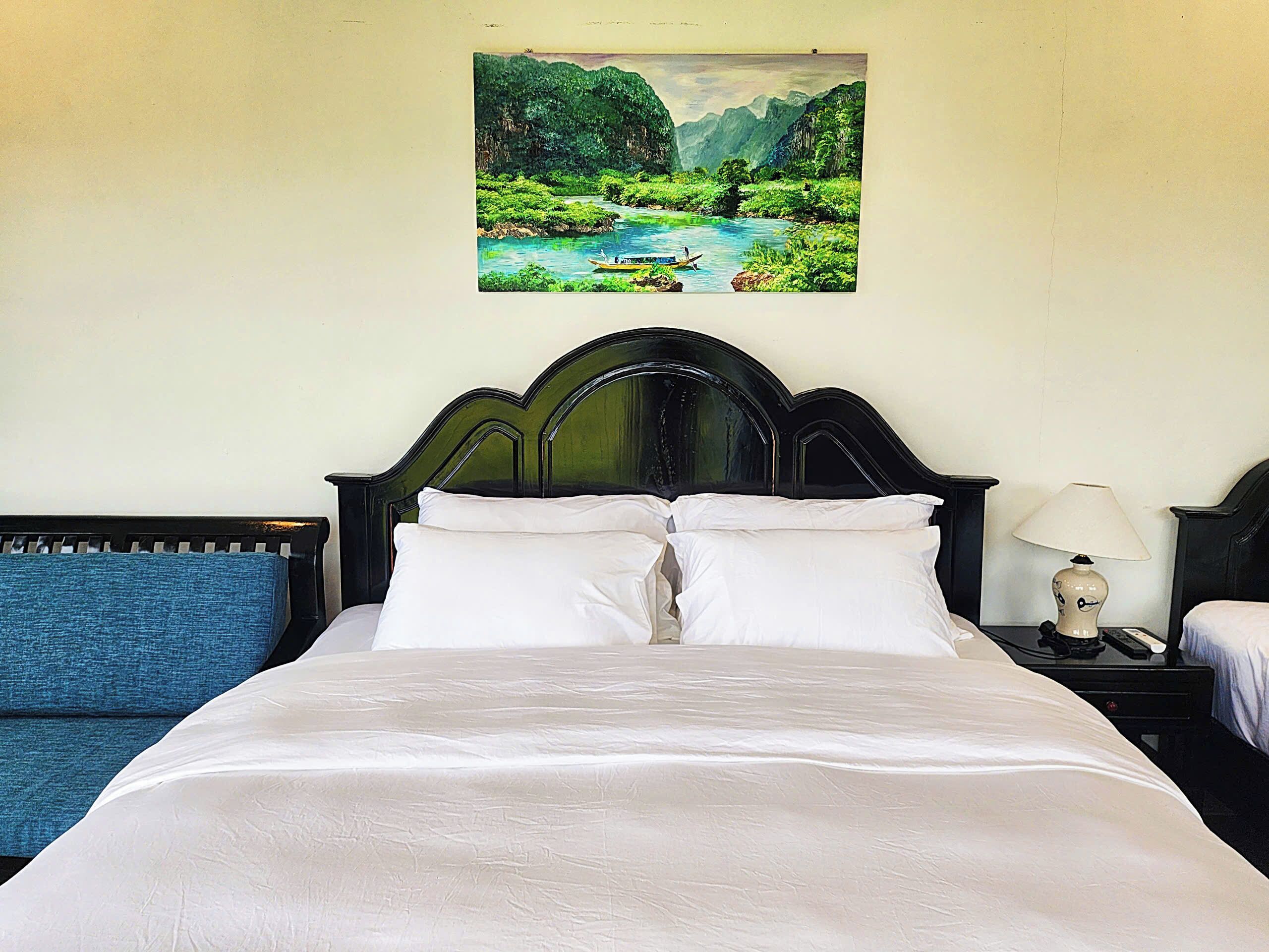 A bed with white bedding, a dark wooden headboard, and a painting of a landscape above it.