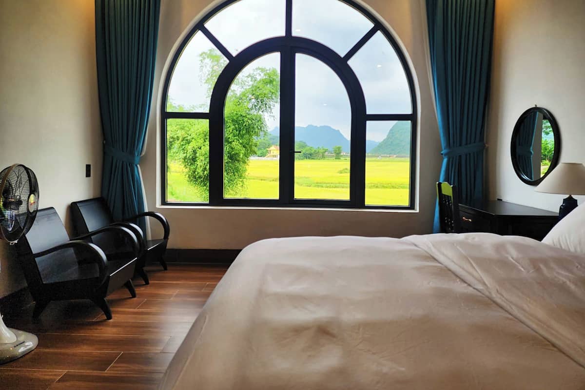 A bedroom with a large arched window, a bed, a desk with a chair, a fan, and a mirror.