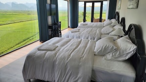 Habitación de hotel con dos camas, un escritorio y un amplio ventanal que ofrece vistas a campos verdes y montañas.