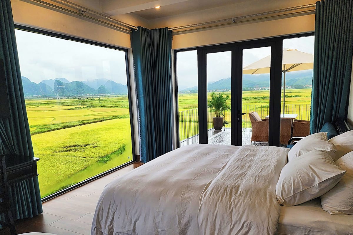 A bedroom with a large bed, a view of a green landscape, and a balcony with a chair.