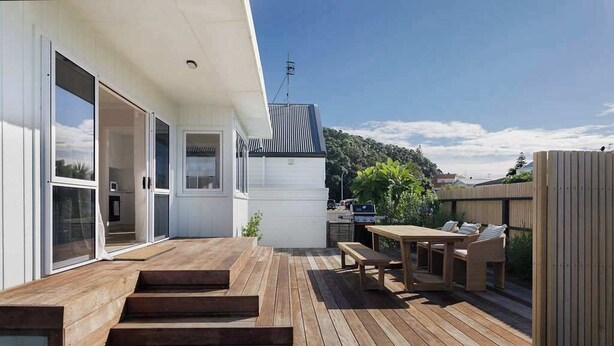 Un terrazzo in legno con tavolo e sedie, un barbecue e una porta scorrevole che dà su un edificio bianco.