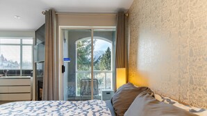 Una camera d'albergo con un letto, una TV, un balcone vista alberi e montagne, e una porta scorrevole.