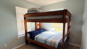 Een stapelbed met houten frame, een raam met zonwering en een lichtschakelaar aan de muur.