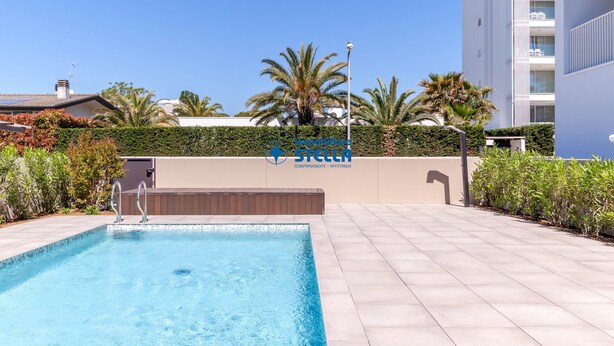 Una piscina con scala in metallo, circondata da solarium piastrellato, palme e un edificio con insegna.