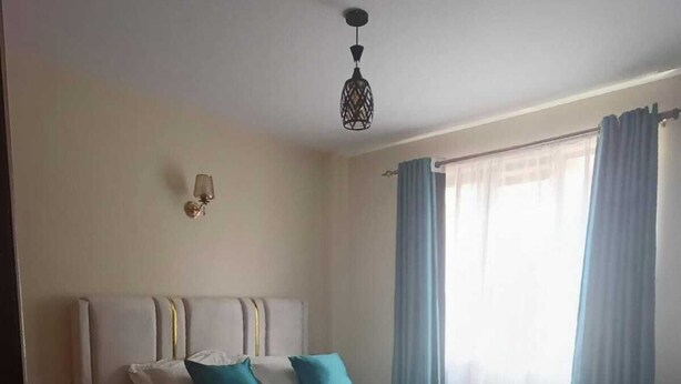 A bedroom with a bed, a wall-mounted light fixture, a window with curtains, and a floor lamp. - A beautiful 2-bedroom unit offering comfort, style, & convenience (Nairobi)