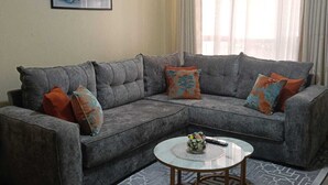 A living room with a grey sectional sofa, a round coffee table, a patterned rug, a painting on the wall, and a window with curtains. - A beautiful 2-bedroom unit offering comfort, style, & convenience (Nairobi)