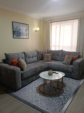 Ein Wohnzimmer mit grauem Ecksofa, einem runden Couchtisch, einem gemusterten Teppich, einem Bild an der Wand und einem Fenster mit Vorhängen.