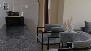 A kitchen with a granite countertop, a microwave, and a coffee maker. There are two grey upholstered chairs and a small table with a vase of flowers. - A beautiful 2-bedroom unit offering comfort, style, & convenience (Nairobi)