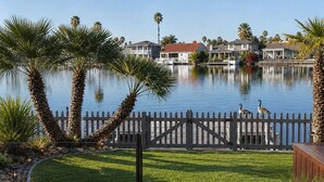 Een pand aan het water met een houten terras, een wit hek en uitzicht op huizen aan de overkant.
