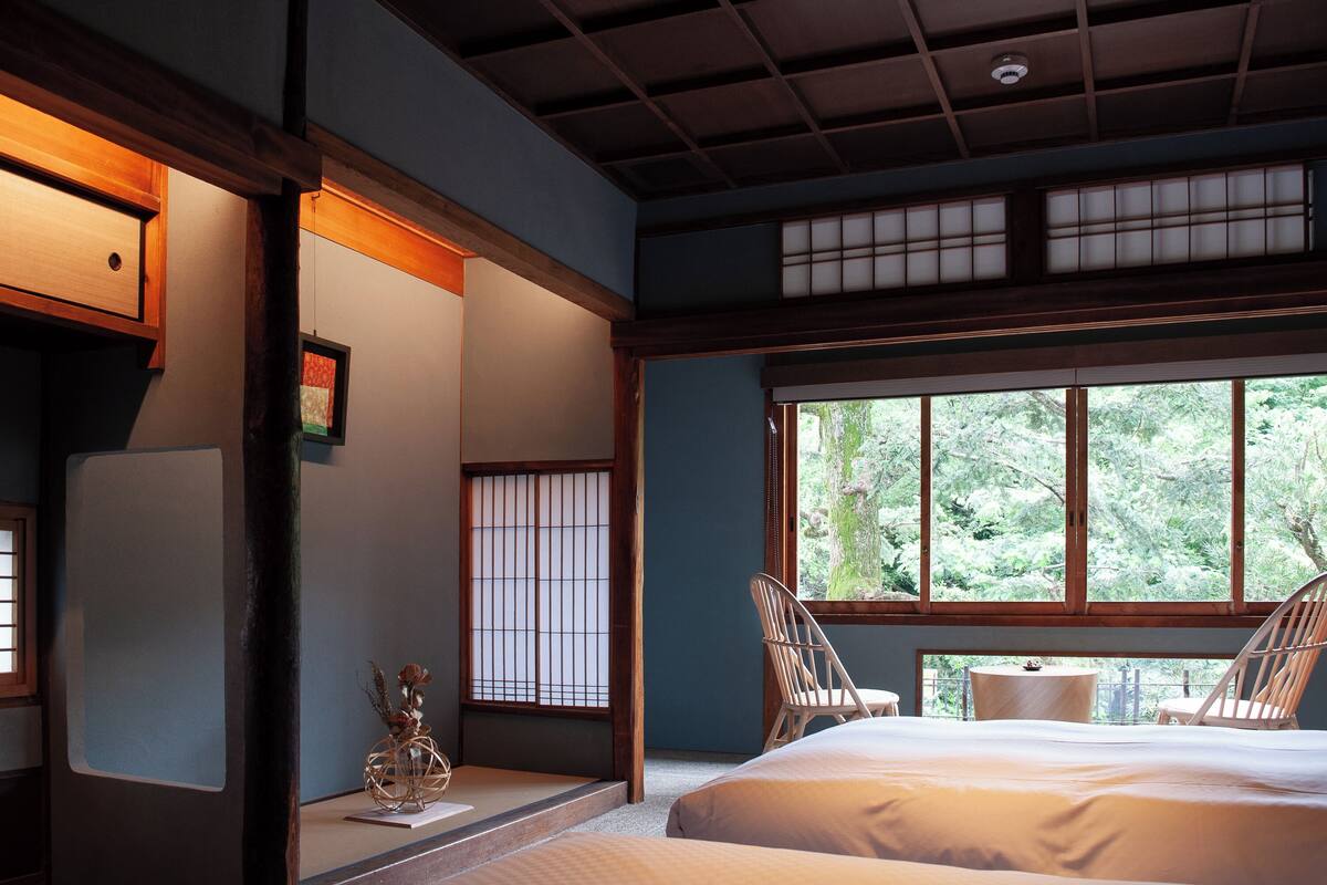Una tradizionale stanza in stile giapponese con pavimento in tatami, un letto, due sedie e un'ampia finestra vista su vegetazione.