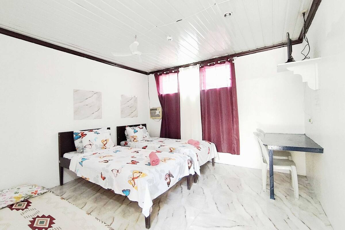 Ein Schlafzimmer mit Bett, Schreibtisch, Stuhl, Deckenventilator und einem Fenster mit Vorhängen.