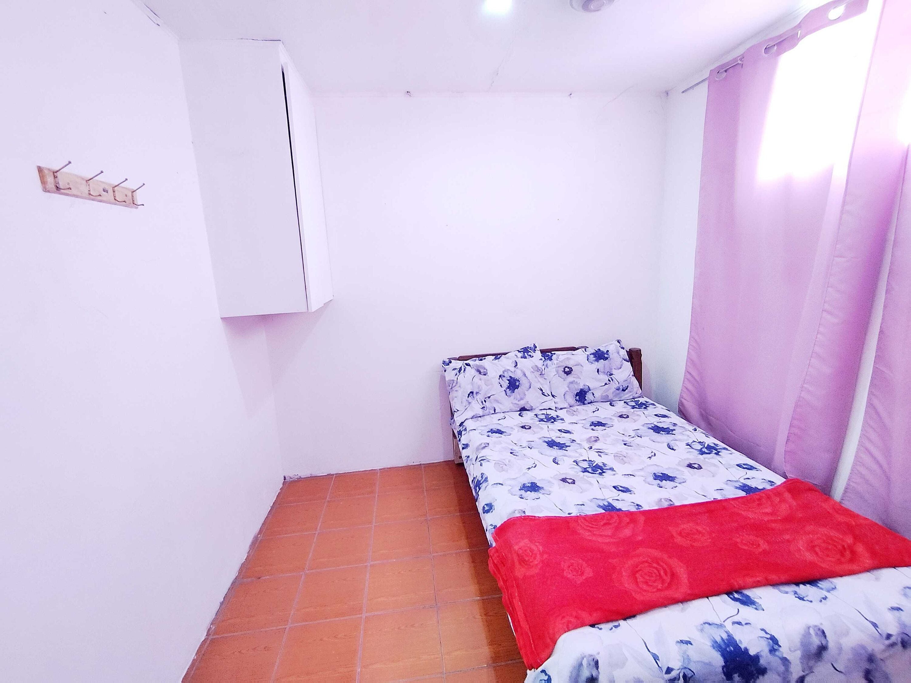 A small, single-bed room with a tiled floor, a white wardrobe, and a window with pink curtains.