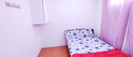 A small, single-bed room with a tiled floor, a white wardrobe, and a window with pink curtains.