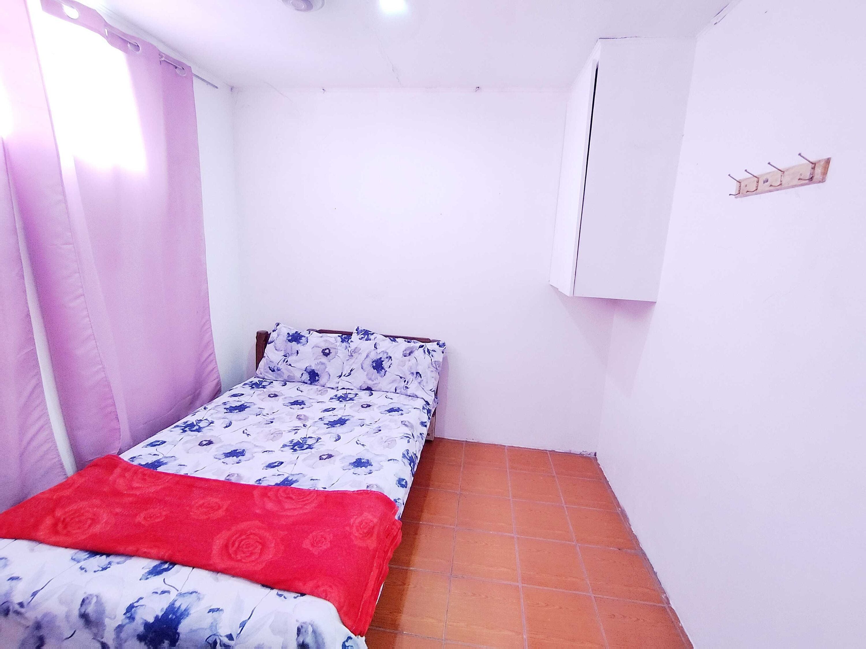 A small, single-bed room with a tiled floor, a bed with floral bedding, a red blanket, and a white wall with a small sign.