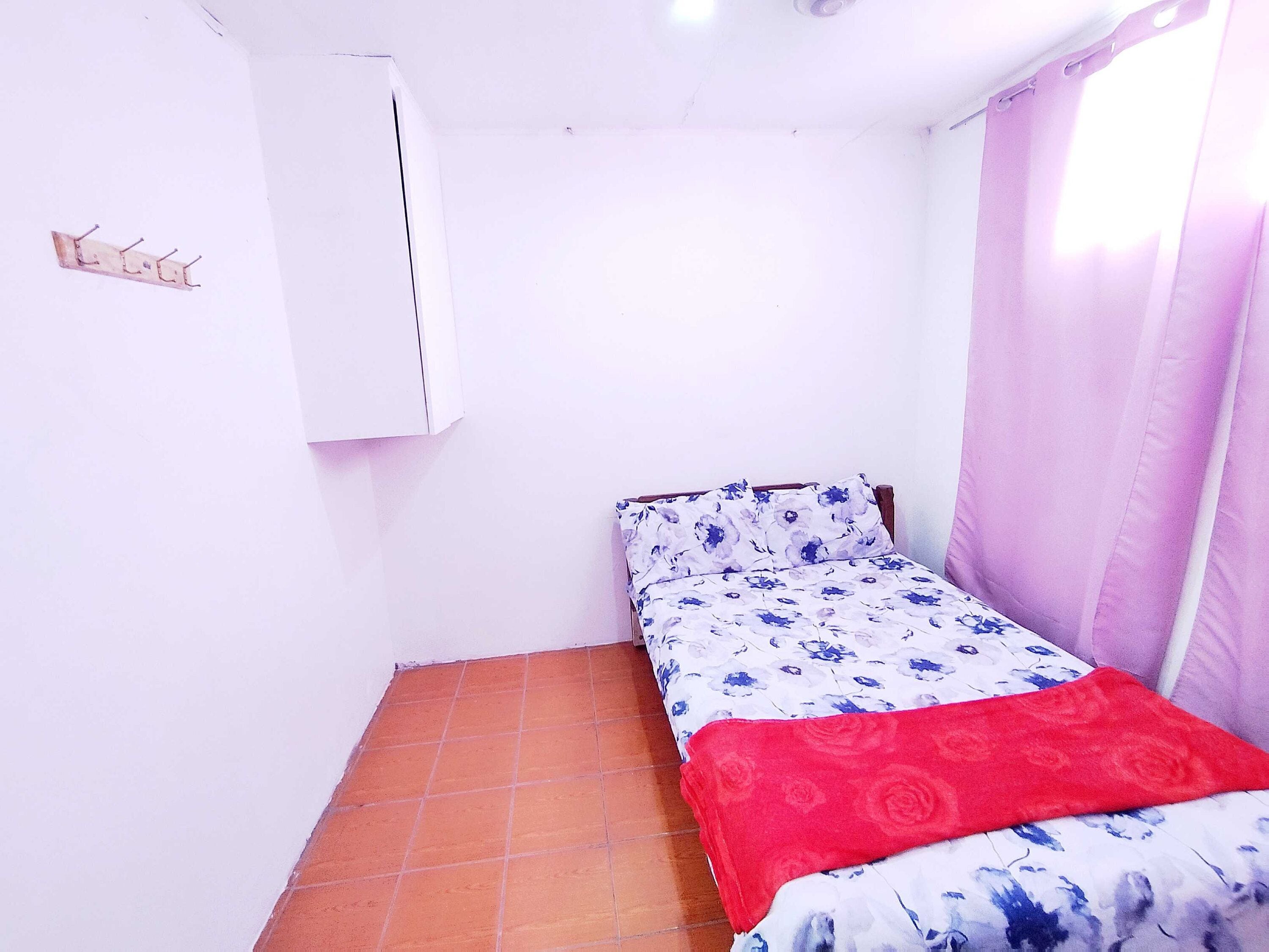 A small room with a bed, a wall-mounted air conditioner, a tiled floor, and a window with curtains.