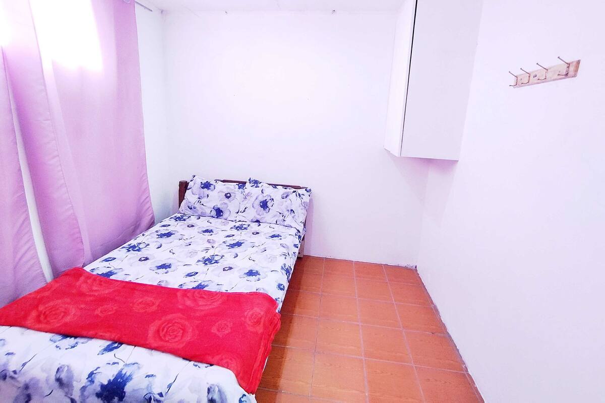 Ein kleines Zimmer mit einem Einzelbett, einer blumigen Tagesdecke, einer roten Decke und einem Fliesenboden.