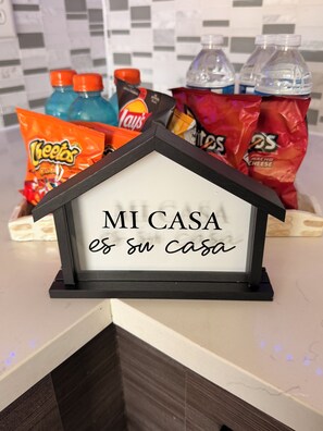 Ein gerahmtes Schild mit der Aufschrift 'MI CASA es su Casa' in einem hausförmigen Rahmen, umgeben von Snacks und Flaschen.