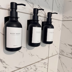 Drei schwarze Flaschen mit Duschgel, Shampoo und Conditioner hängen an einer Wand mit Marmorstruktur.