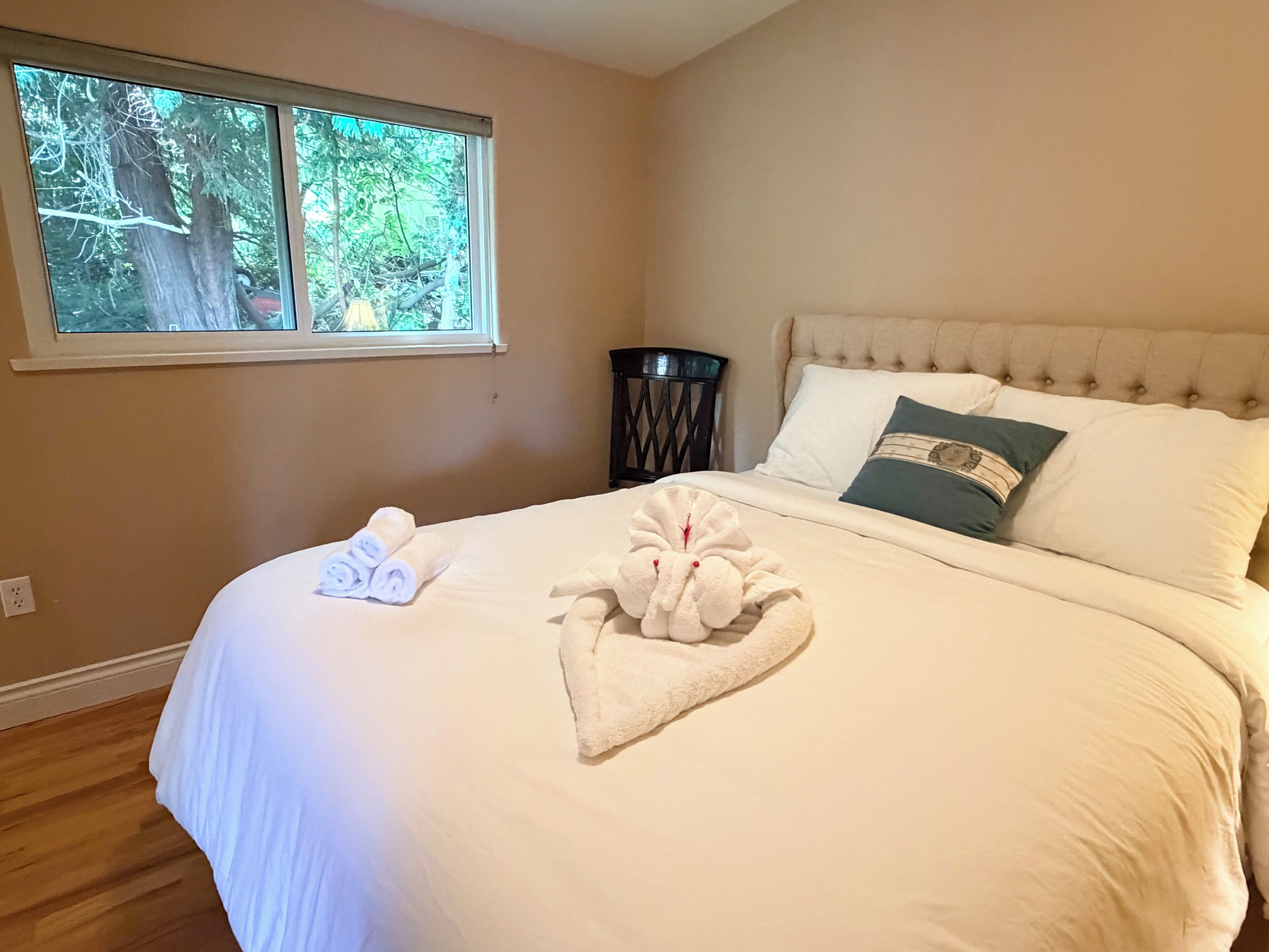 Un lit bien fait, avec des serviettes pliées et un coussin décoratif, une fenêtre donnant sur des arbres et une chaise sombre placée à côté du lit.