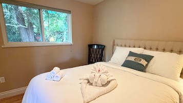 Un lit bien fait, avec des serviettes pliées et un coussin décoratif, une fenêtre donnant sur des arbres et une chaise sombre placée à côté du lit.