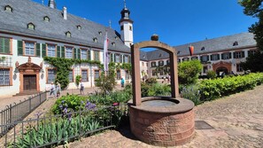 A courtyard with a well-maintained garden, a stone well, and traditional buildings with a church steeple. - MainZeit Seligenstadt - Your half-timbered dream in Seligenstadt (Seligenstadt)