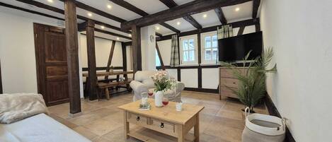 Ein gemütliches Zimmer mit freiliegenden Holzbalken, einer Couch, einem Couchtisch mit Büchern und Blumen, einem Fernseher und einem Fenster mit Vorhängen.