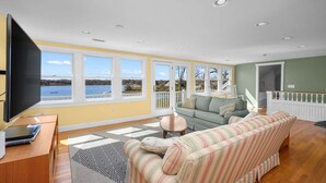 A living room with a large window, a sofa, a TV, and a wooden desk. - Waterfront Chatham Escape, Pet Friendly, Pond Views & Beach Nearby (Chatham)