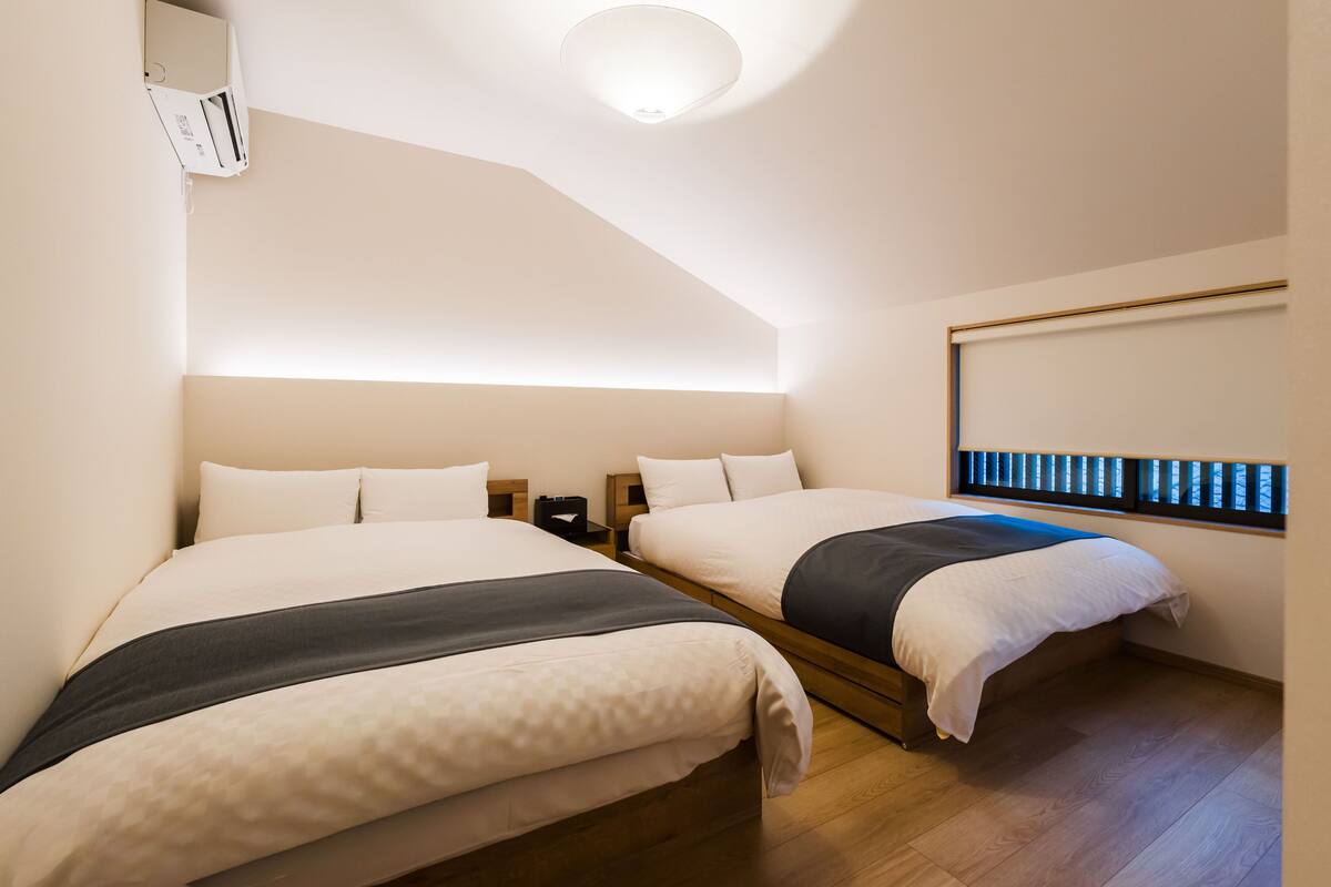 Ein Hotelzimmer mit zwei Betten, einer Deckenlampe, einem Fenster mit Jalousien und Holzboden.