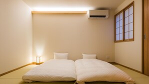 Una habitación con suelo de tatami, cama con ropa blanca, aire acondicionado, ventana con paneles shoji y mesita de noche con lámpara.