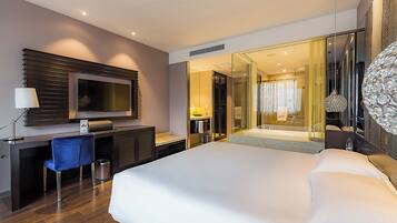 A modern hotel room with a large bed, a desk with a chair, a TV, and a glass-enclosed bathroom.